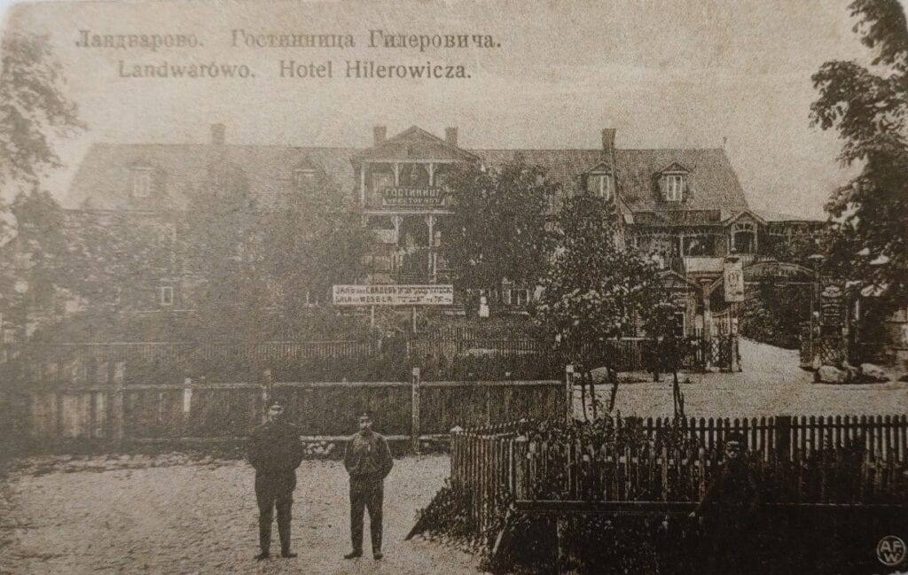 Sepia postcard of a large wooden hotel behind a fence, two men standing in the foreground